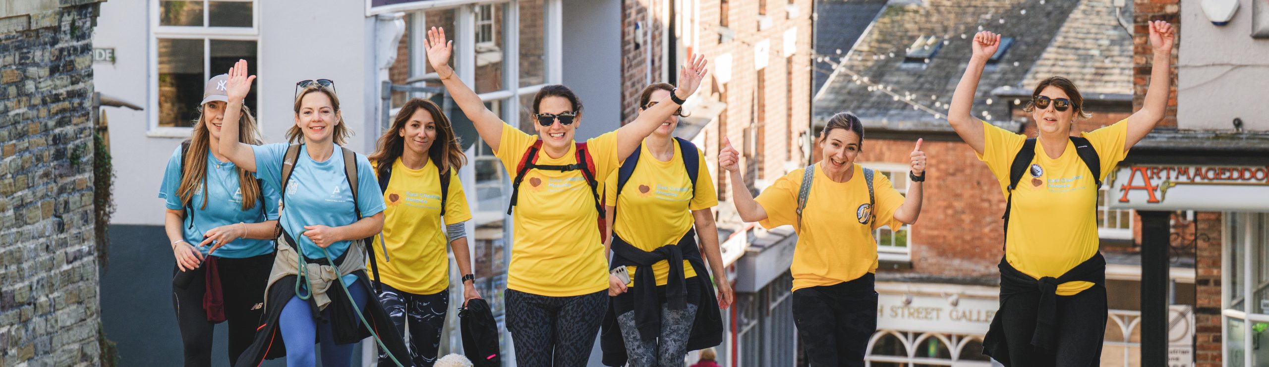 A group of smiling women wearing matching yellow and blue T-shirts walk outdoors, raising their arms in celebration as they participate in a Live Your Life group walk or event on a sunny day in a town.