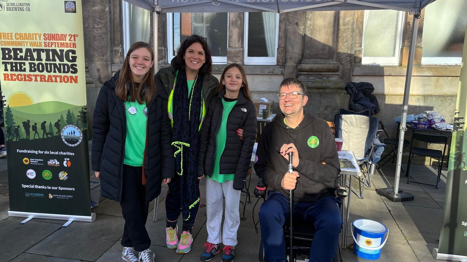 Four people, three standing and one seated in a mobility scooter, smile under a tent at an outdoor event. Banners behind them promote "Beating the Bounds Registration"—a charity event inspiring all to live your life fully. Four people, three standing and one seated in a mobility scooter, smile under a tent at an outdoor event. Banners behind them promote "Beating the Bounds Registration"—a charity event inspiring all to live your life fully.