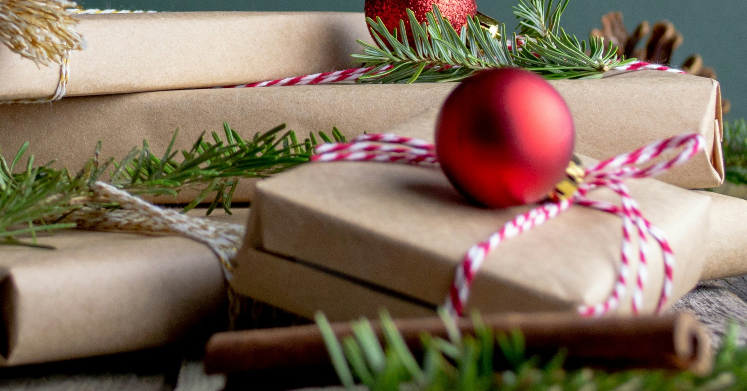 Several gifts wrapped in brown paper and tied with red-and-white string are decorated with pine branches and red Christmas ornaments, celebrating the spirit of the 12 Days with a cinnamon stick in the foreground.