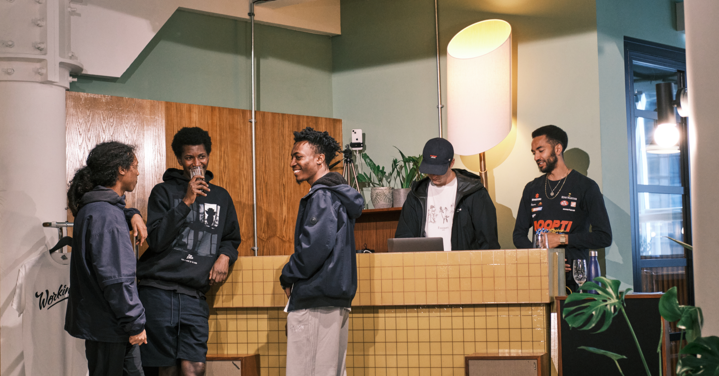 Five people stand and chat casually near a tiled counter in a modern, warmly lit indoor space with wood paneling, plants, and a large lamp. The relaxed group shares stories of falling upwards as they enjoy their engaging conversation.