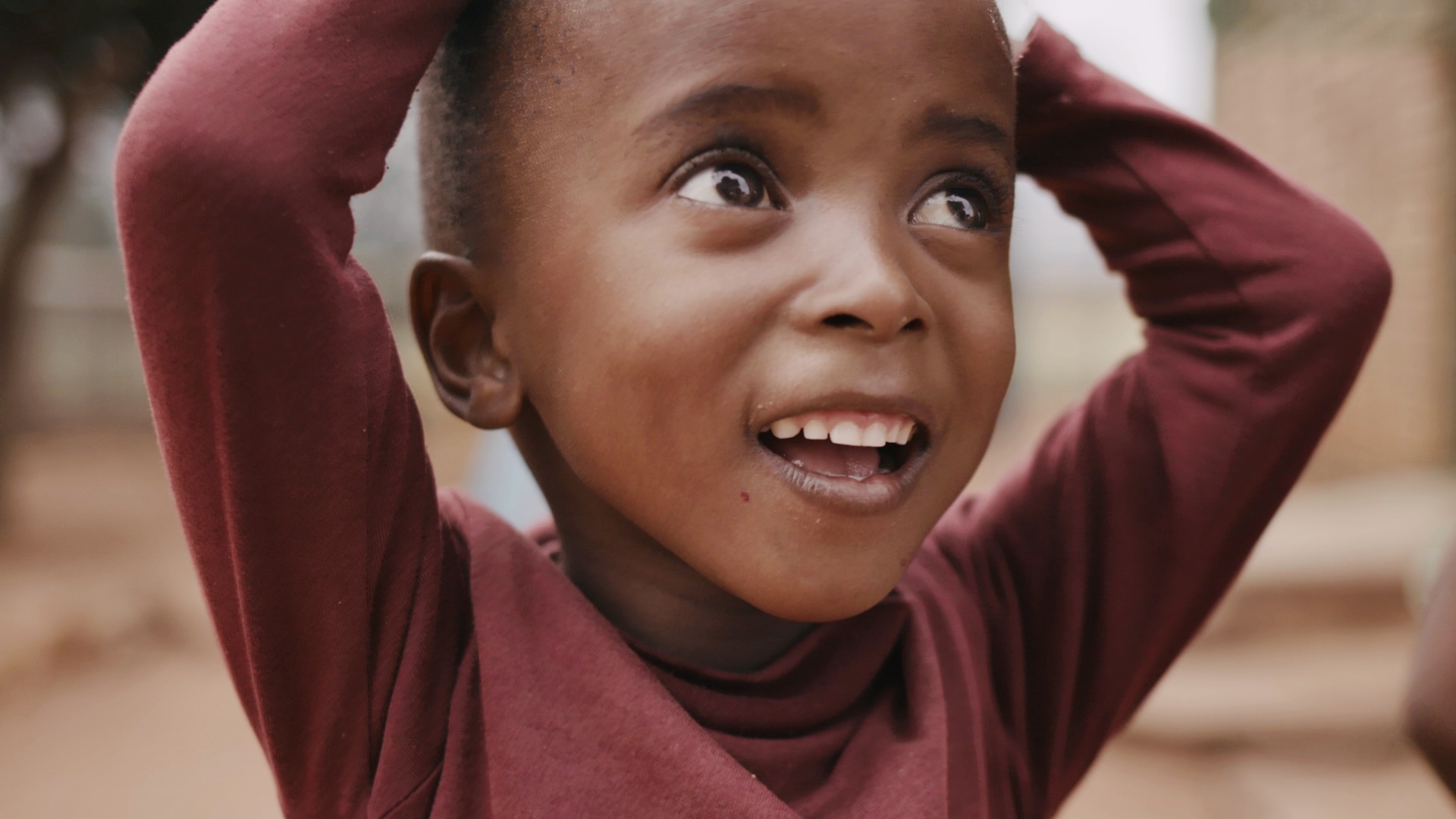 A young child with a joyful expression looks upwards, perhaps wondering about the unpredictability of outer space. They are wearing a maroon top and have their hands resting on their head. The background is softly blurred, suggesting an outdoor setting. The child's eyes are wide open, and they have a small gap between their front teeth.