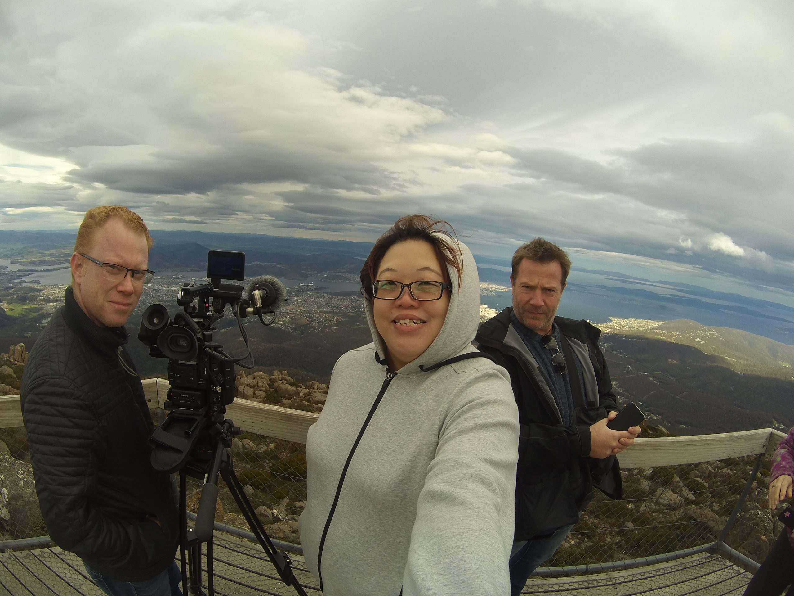 Three people stand on a wooden platform with a scenic view of mountains and water in the background. One person takes a selfie while others stand near a professional video camera. The sky is cloudy.