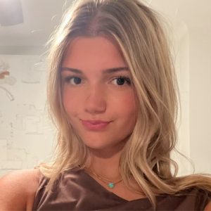 A young woman with long, blonde hair and a slight smile poses for a selfie indoors. She is wearing a brown top and two delicate necklaces, one with a turquoise pendant. The background is softly lit and out of focus.