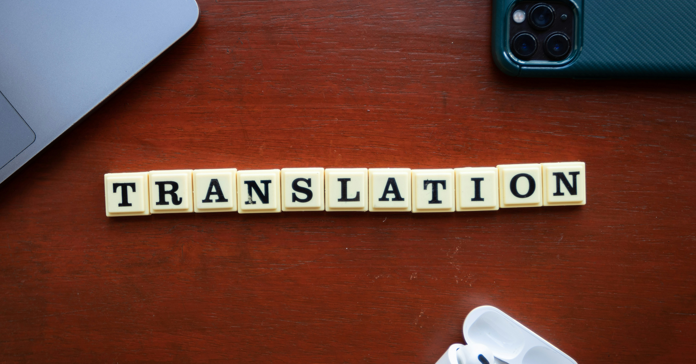 Yellow letter tiles spelling “TRANSLATION” are arranged in a row on a wooden table, surrounded by part of a laptop, a green smartphone, and a white wireless earbud case—highlighting how easy words can bridge cultural disconnect.