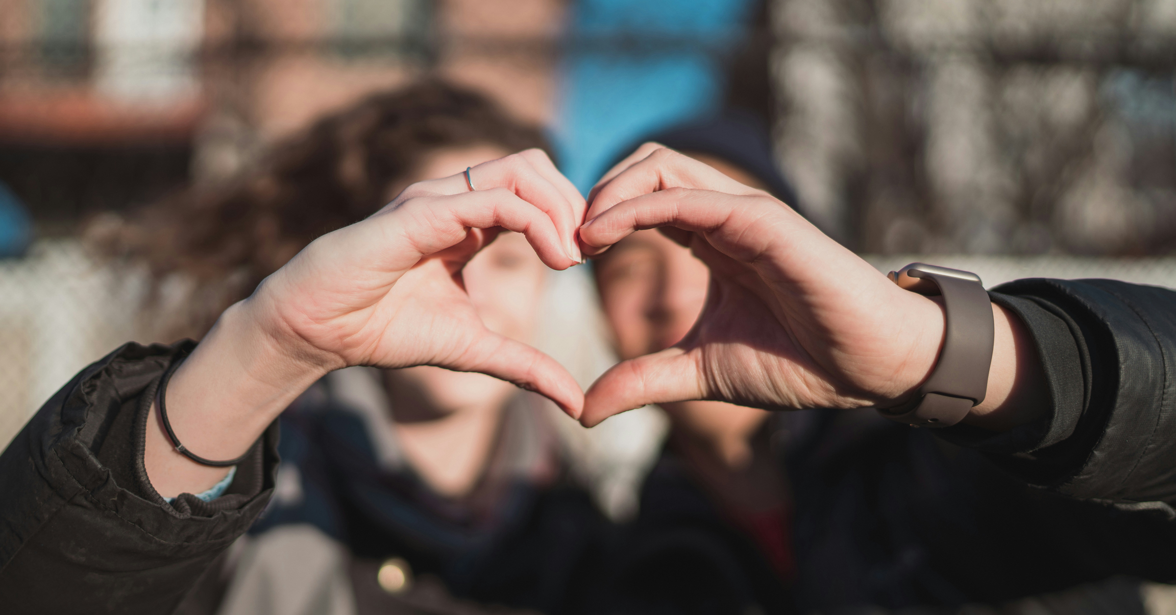 Two people forming a heart shape with their hands in the foreground, with their faces blurred in the background, outdoors on a sunny day.