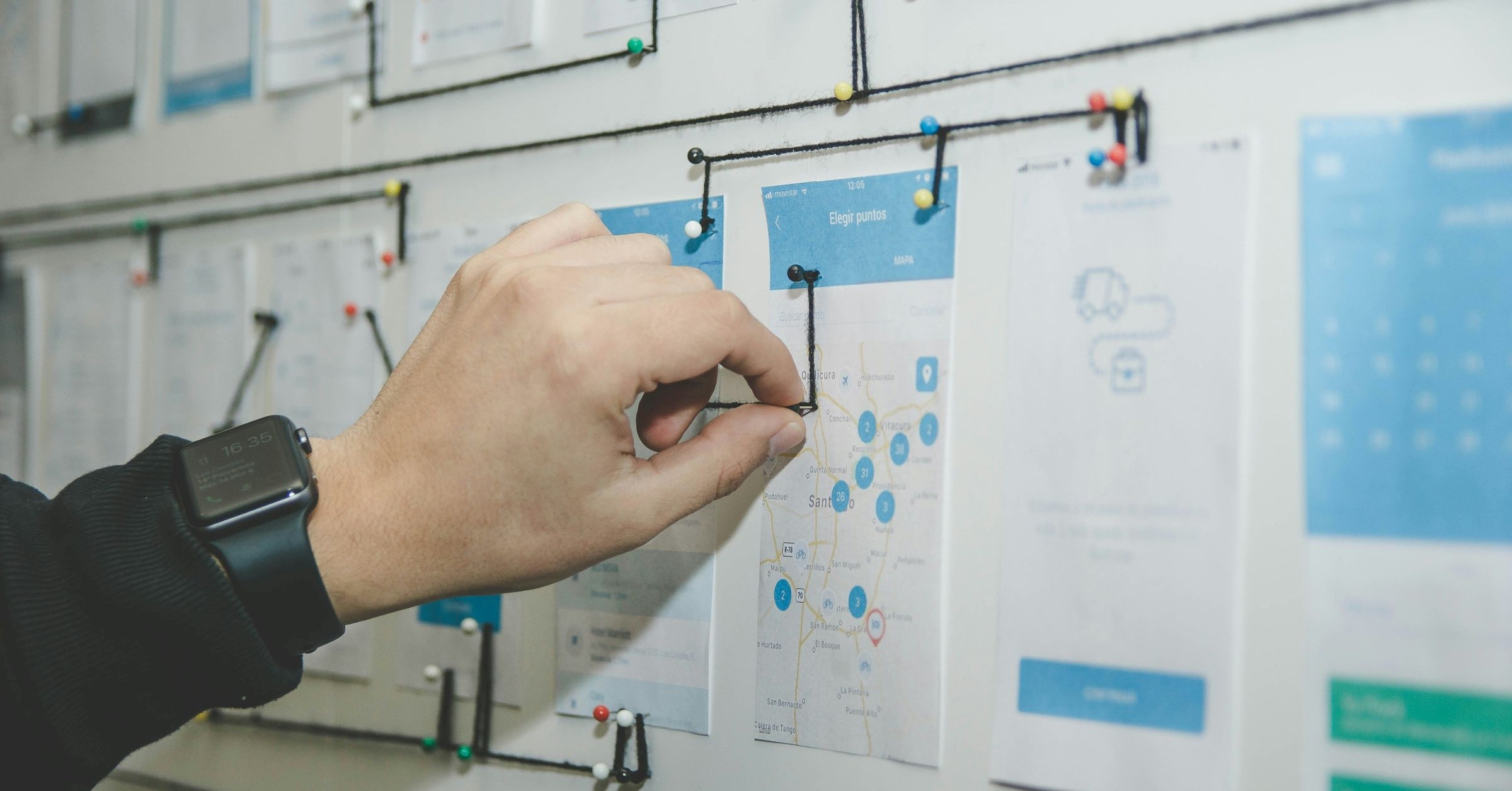 A person wearing a smartwatch pins a string to a board covered with app interface designs and diagrams, mapping out a project to streamline content creation.