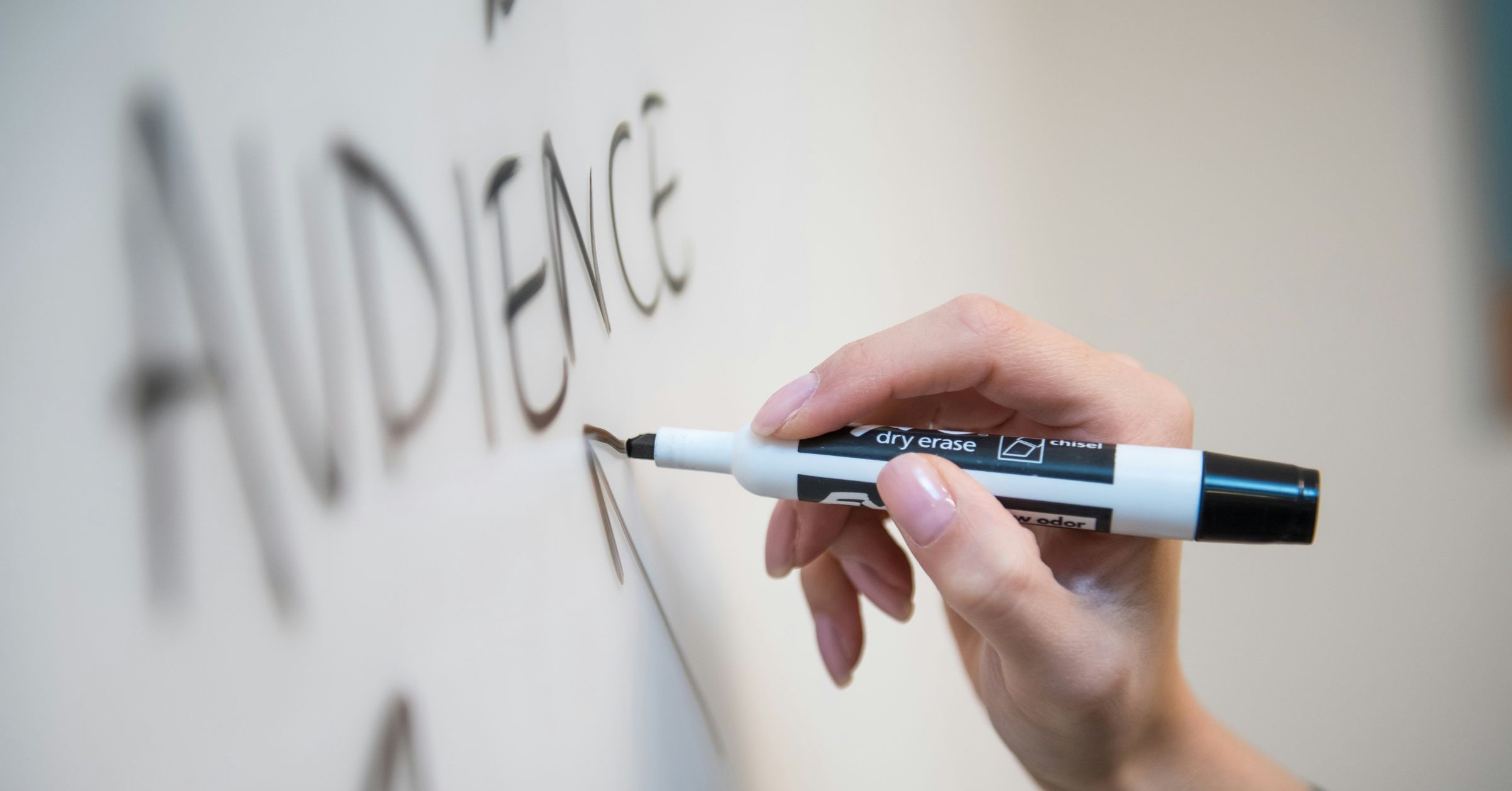 A hand holding a black dry-wipe marker writes the word "AUDIENCE" in large letters on a whiteboard, illustrating the vital role of audience insight in moving from awareness to decision along the customer journey.