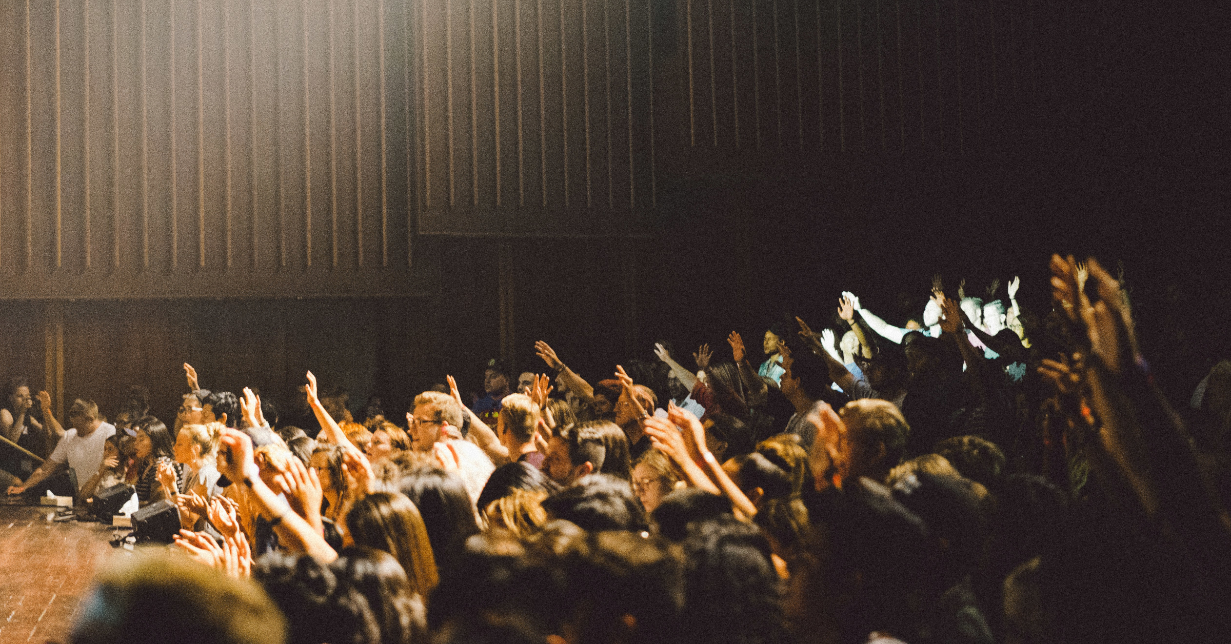 A large audience sits in a dimly lit auditorium, with many people raising their hands—perhaps eager to reveal a hidden talent—as they face the stage on the left. Light highlights some, while others remain in shadow.