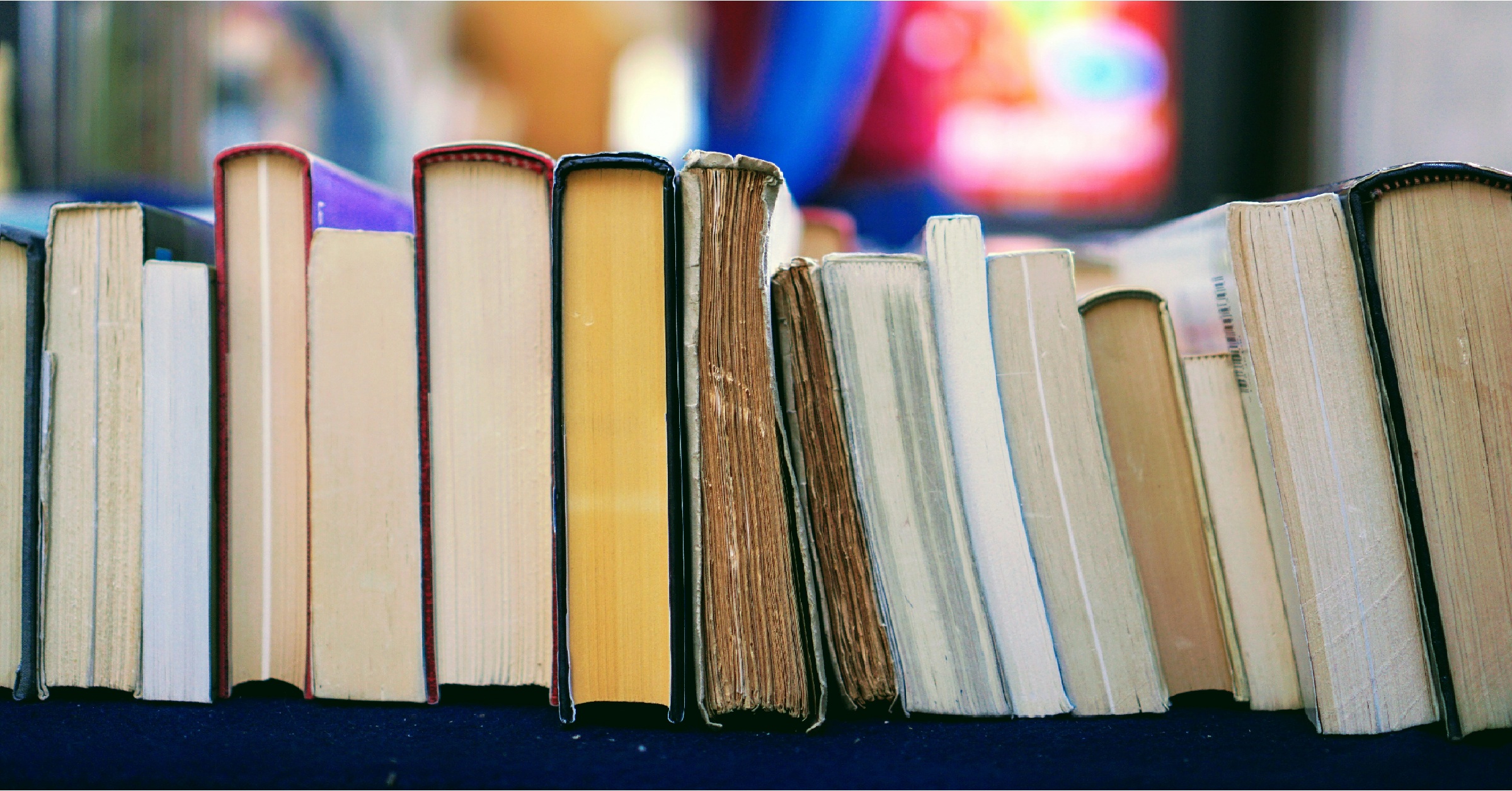 A row of books standing upright with their spines and pages facing forward, showing a variety of colors and thicknesses, on a dark blue surface with a blurred, colorful background.