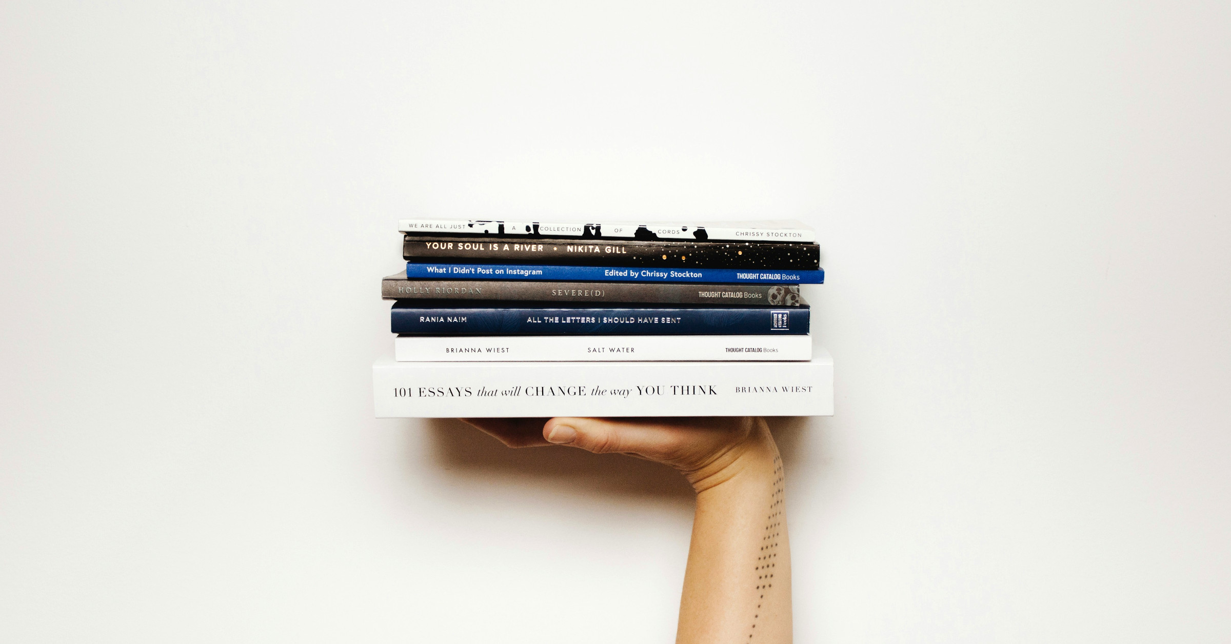 A hand with a dotted-line tattoo on the forearm holds a stack of seven books against a plain white background. The books are stacked horizontally and viewed from the side.