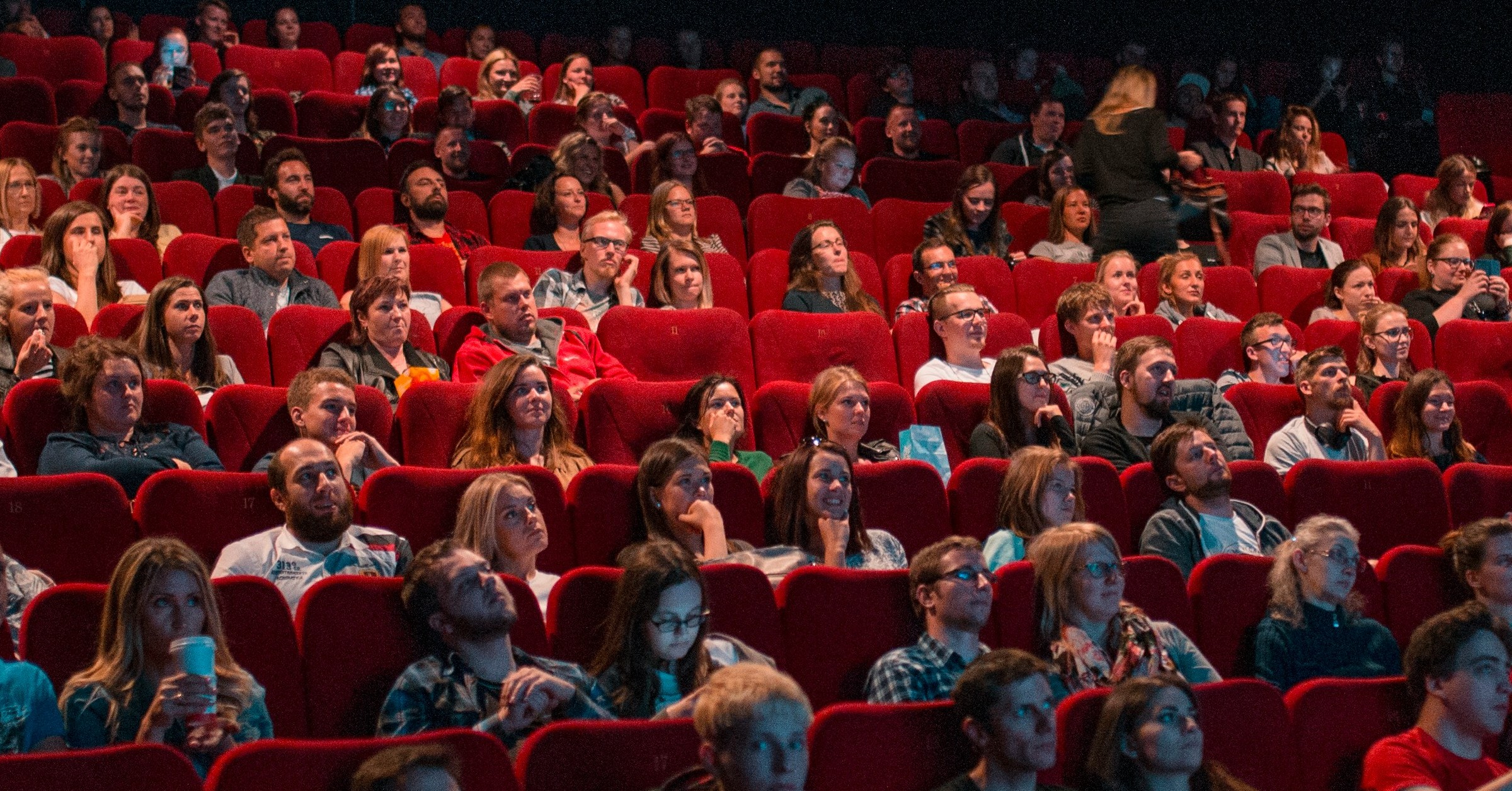 A large audience of cinematic universe advocates sits in red theatre seats, watching an event or film. Most people are facing forward and appear attentive, while one person is standing and moving along the aisle.
