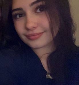A young woman with long dark hair, dark eyes, and subtle make-up smiles softly at the camera. She is wearing a dark top and a necklace with a pendant. The background is softly lit and neutral.