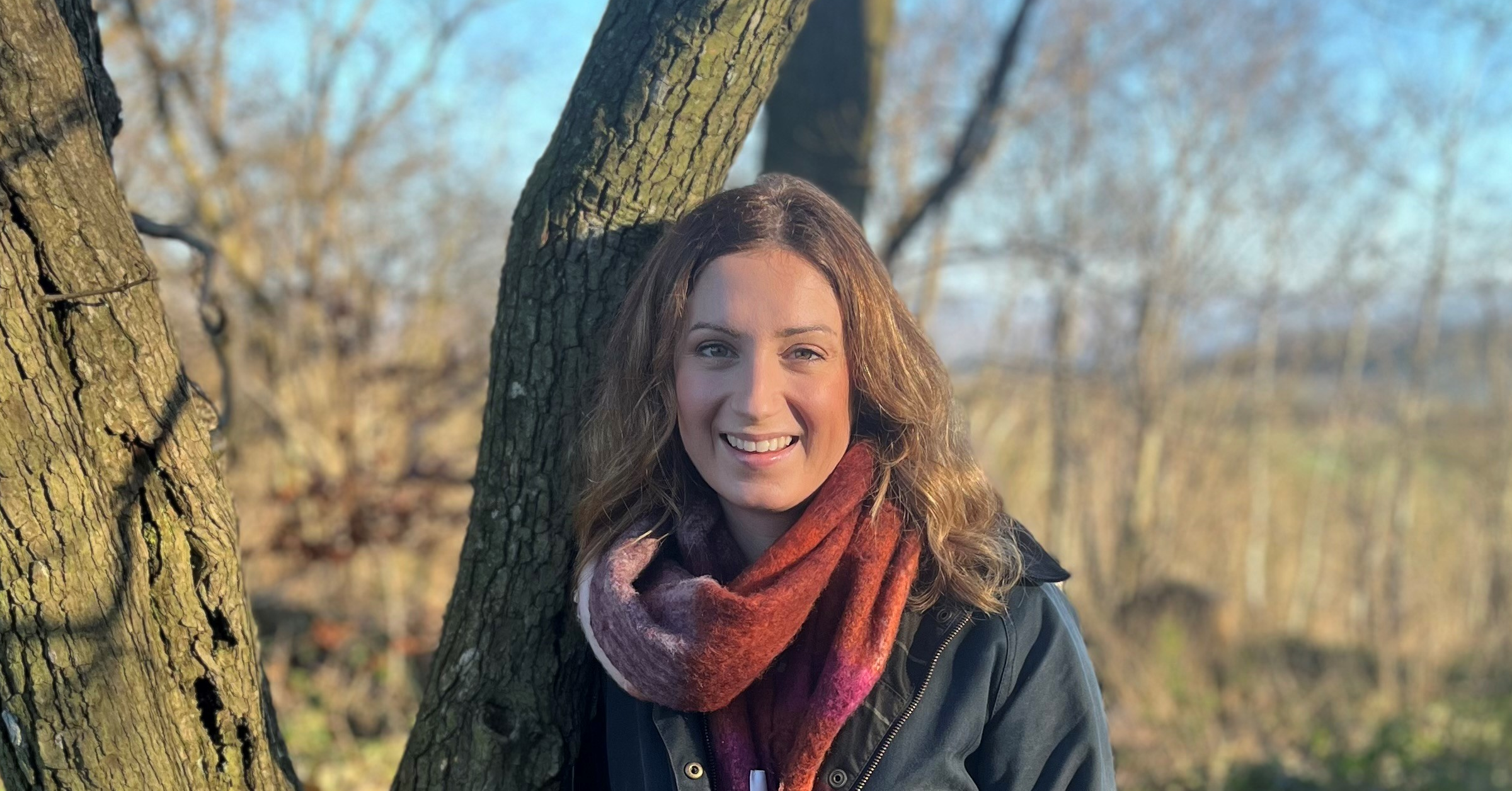 A woman with wavy brown hair and a red scarf smiles while standing outdoors by a tree on a sunny day, with a blurred background of leafless trees and blue sky.
