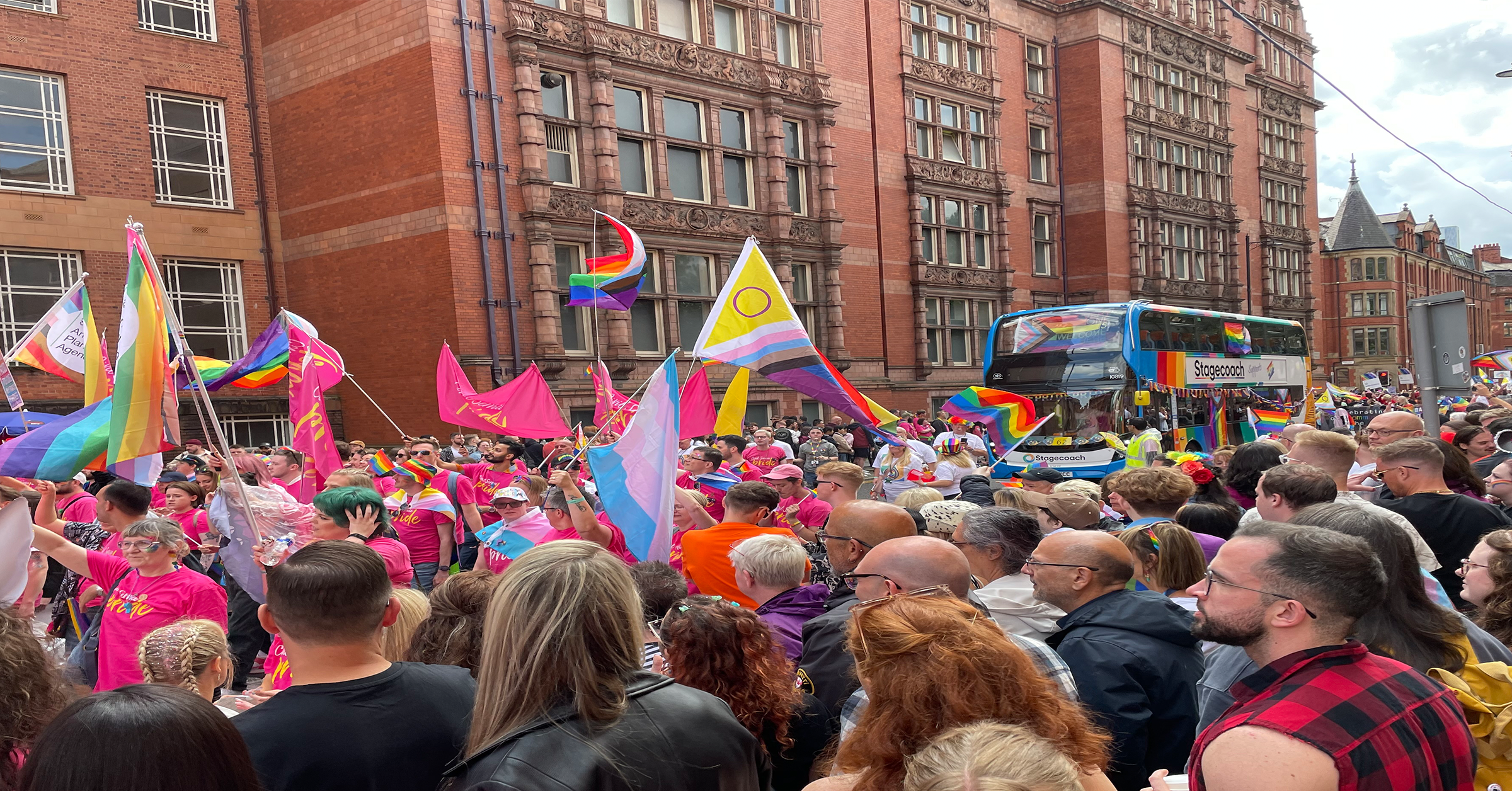 A large crowd gathers at a Pride Month parade, waving rainbow, trans, and other LGBTQ+ flags on a city street lined with red brick buildings. A double-decker bus with rainbow decorations is visible in the background for this Pride Month article.