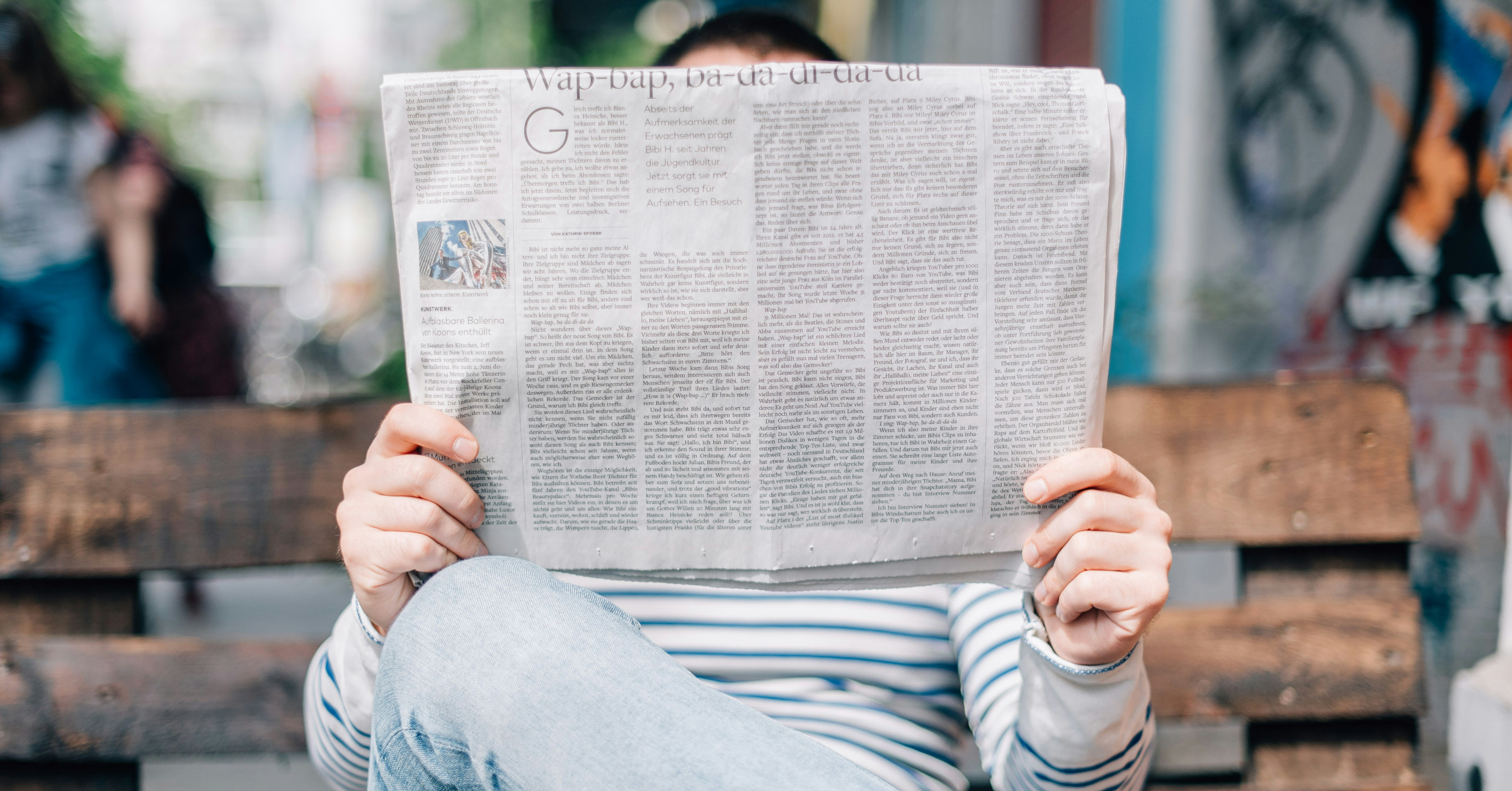 A person wearing blue jeans and a striped shirt sits on a wooden bench, holding and reading a large newspaper that covers their face. The background is blurred.