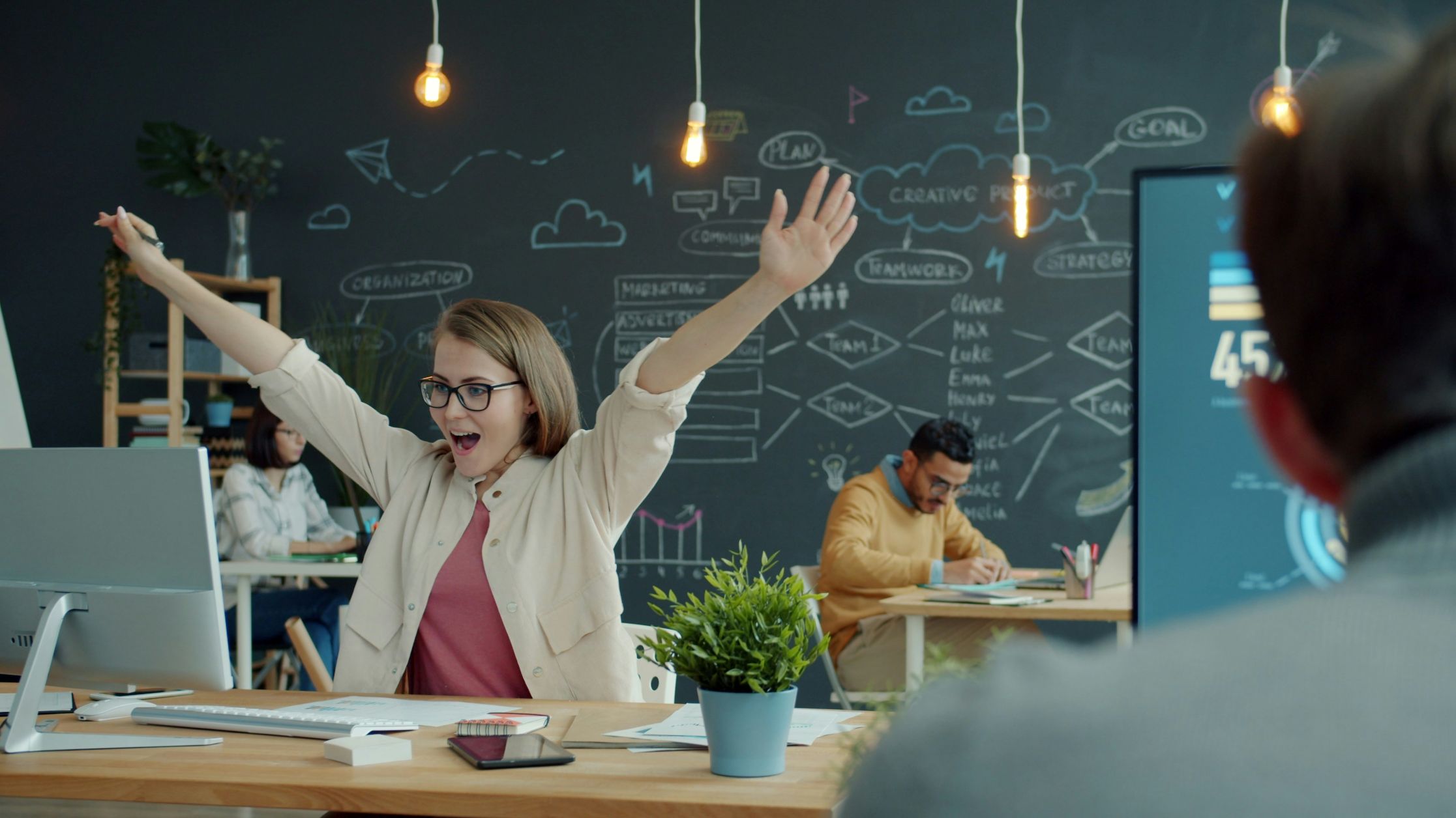 A woman in glasses excitedly raises her arms at her desk in a modern office, celebrating the success of an employee advocacy program. Behind her, coworkers work at desks; a chalkboard wall with charts and doodles is visible. A plant and computer sit on her desk.