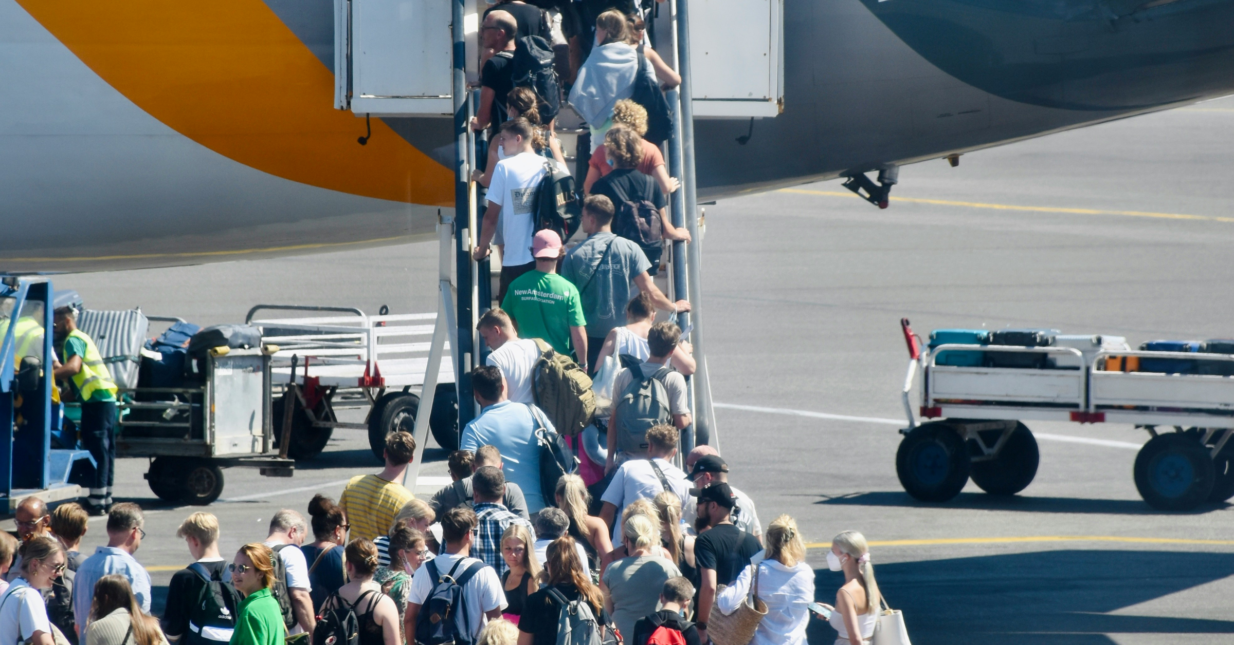 A large group of travellers waits in queue and boards an aeroplane via a stairway on the tarmac, with luggage trolleys and airport staff visible nearby.