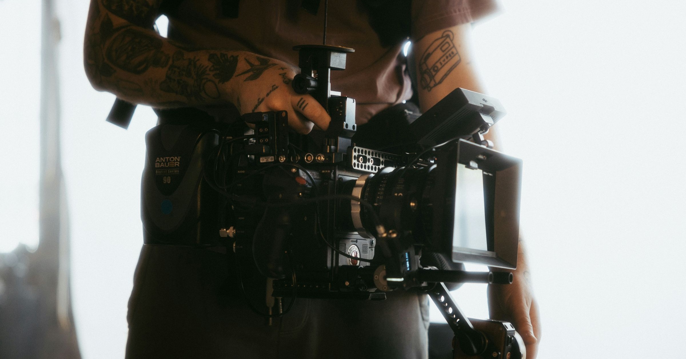 A tattooed video producer holds a professional camera rig, preparing to film. The bright, mostly white background emphasizes the video production equipment.