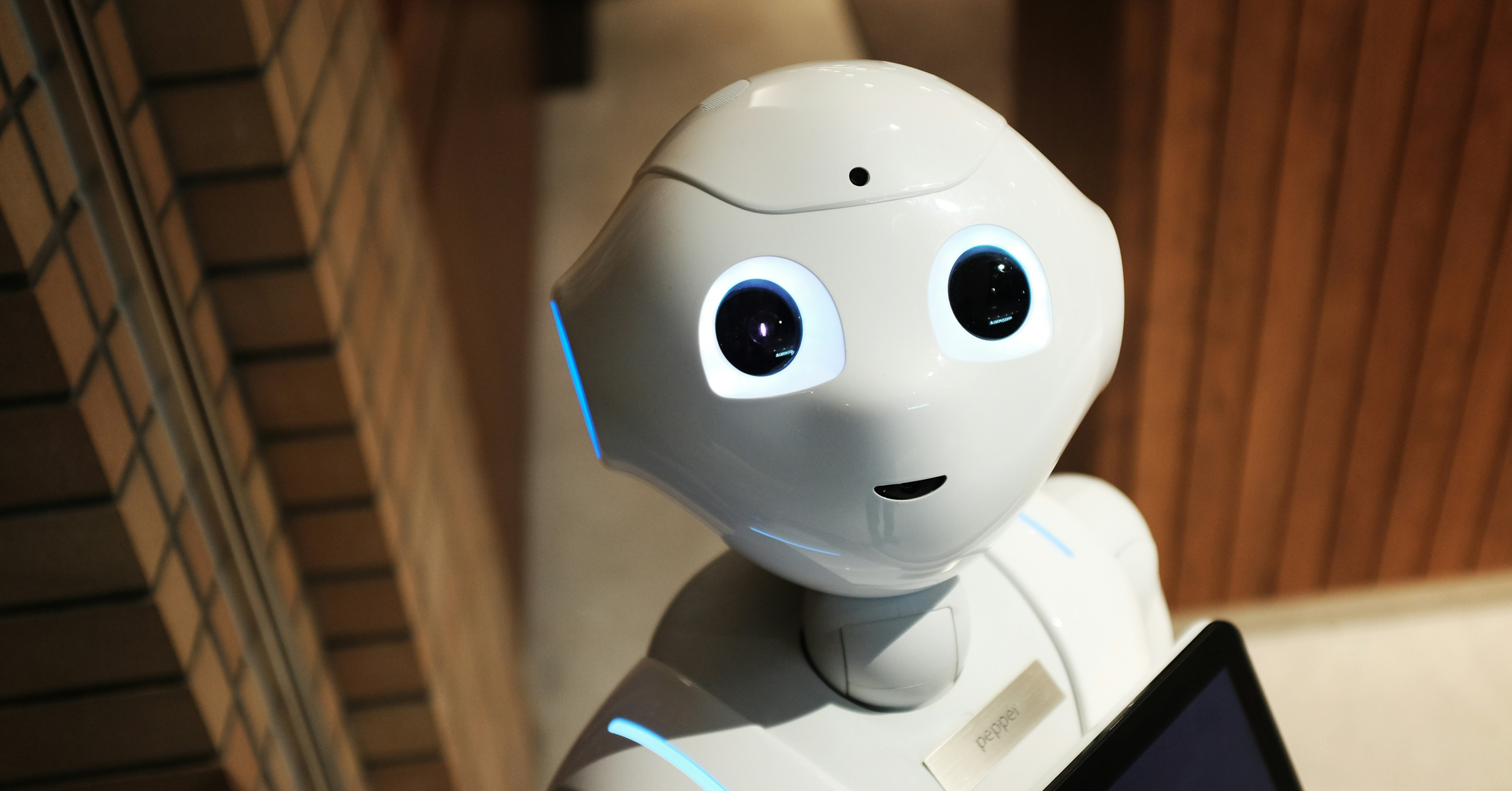 A white humanoid robot with large, round black eyes and a friendly expression looks up, ready to show off the latest tech. The background features a brick wall on the left and wooden panels on the right as it holds a black tablet for audiences.