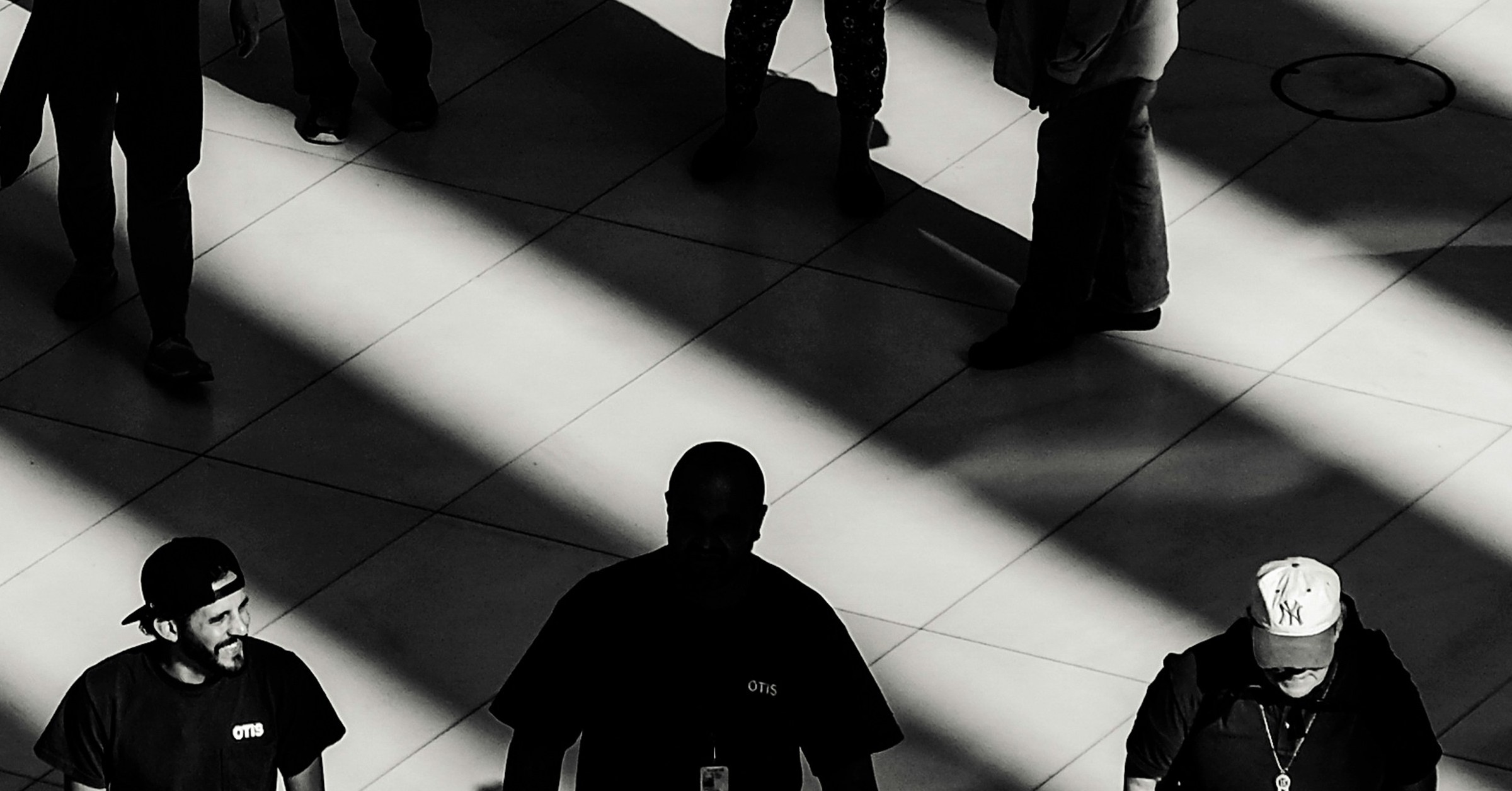 Black and white photo of people walking on a tiled floor, casting long shadows. Like a perfect case study, light and shadow interact to create a striking geometric pattern among figures in baseball caps and dark shirts.