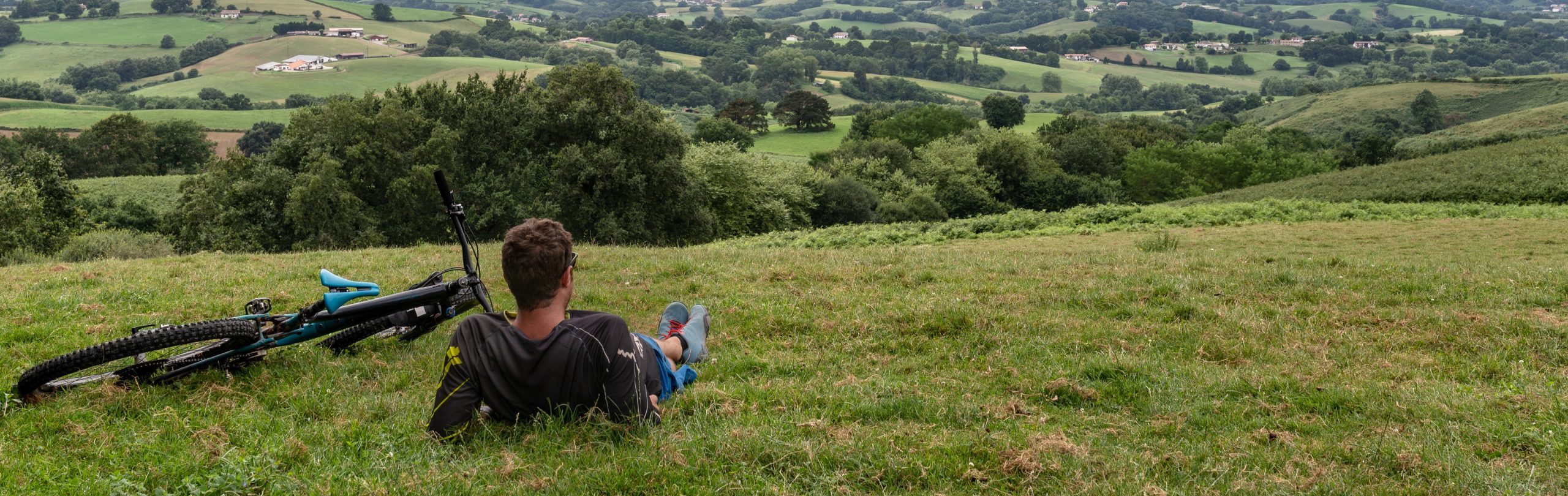 A person relaxes on a grassy hillside next to a mountain bike, looking out over green fields and rolling hills as Dani Bobe and their team ride through the distant mountains under a cloudy sky.