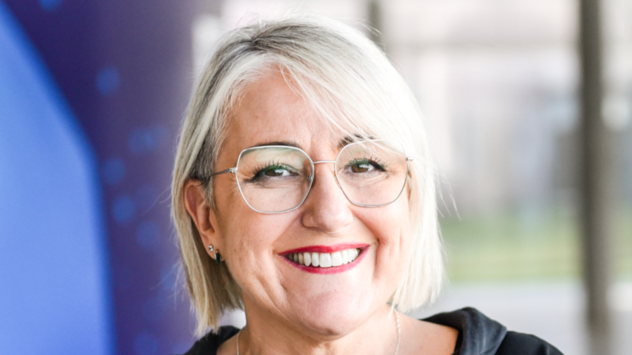 A woman with short blond hair and glasses smiles at the camera. She is wearing a dark top and has red lipstick. The background is blurred with blue and neutral tones.