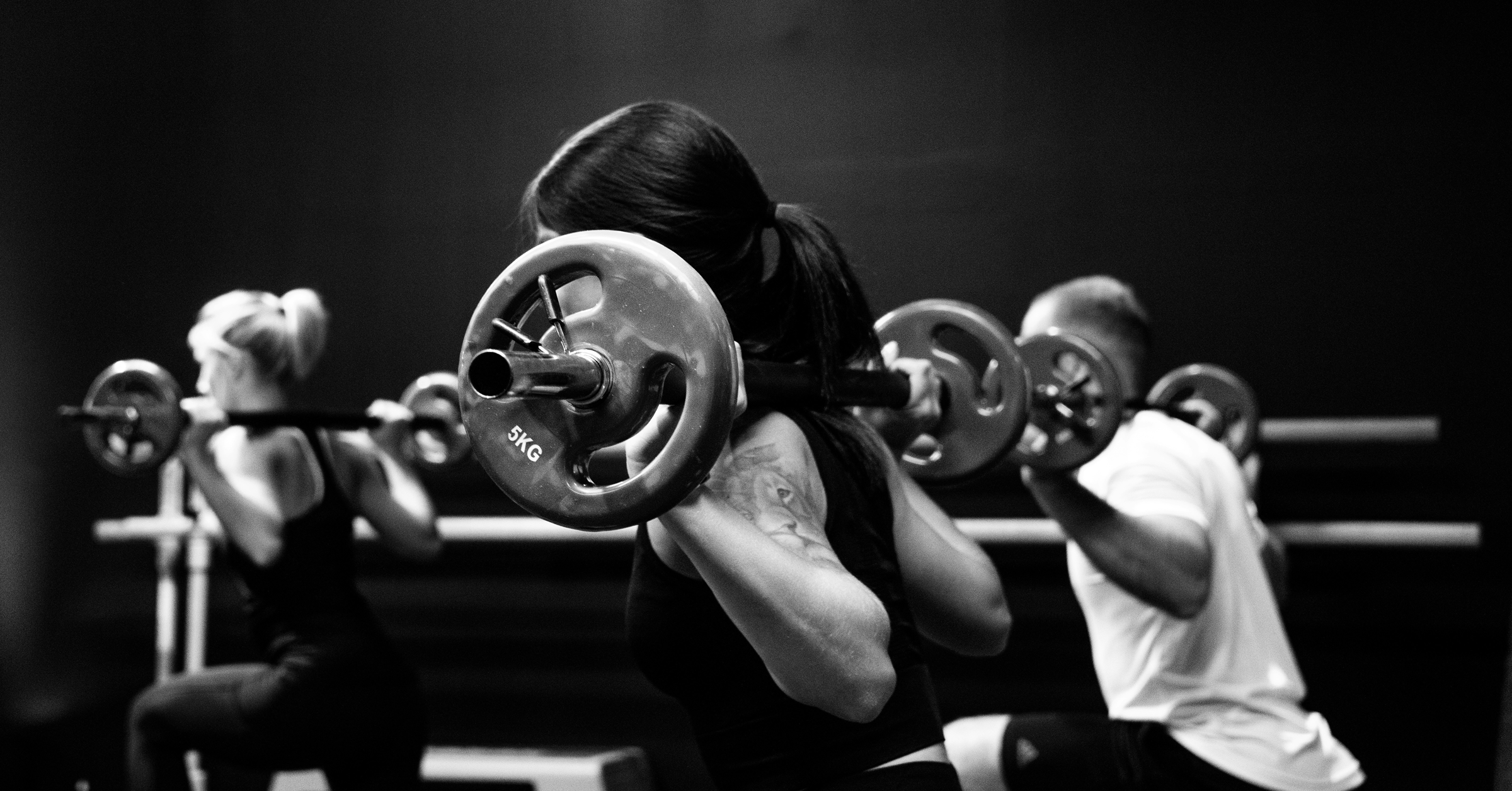 Three people in workout clothes lift barbells on their shoulders whilst performing squats in a gym. The black and white image captures stronger stories of strength and focus during a group exercise session.