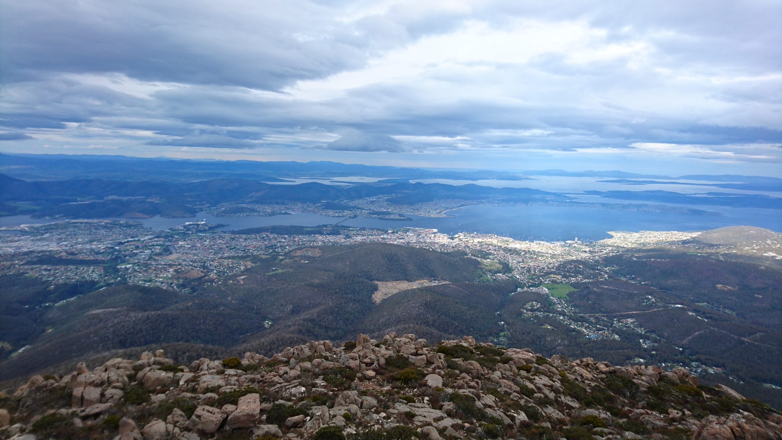 A panoramic view from a mountaintop overlooking a city surrounded by water. Rolling hills and a vast sky with scattered clouds complete the landscape. Rocks and sparse vegetation are visible in the foreground.