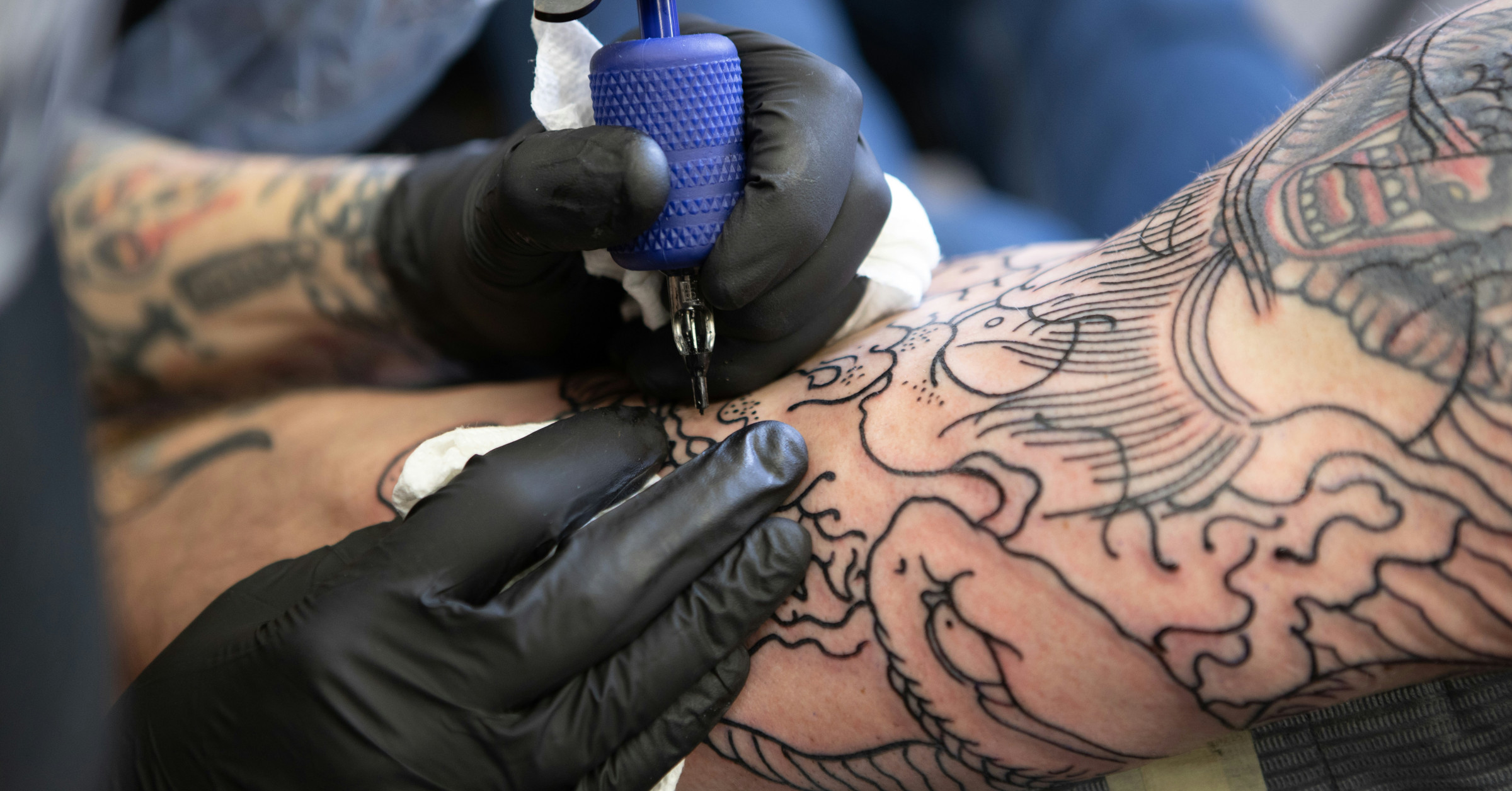 A tattoo artist wearing black gloves uses a tattoo machine to ink an intricate line design on a person's arm, which is already covered with various detailed black line tattoos.