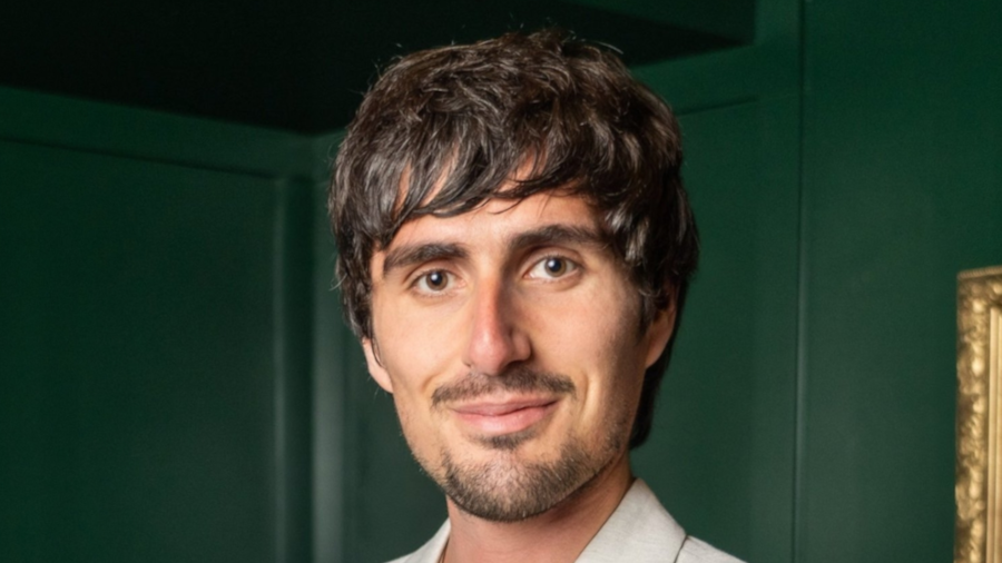 A man with short brown hair and a trimmed beard, wearing a light-colored jacket and smiling, stands in front of a dark green wall with part of a gold-framed picture visible on the right.